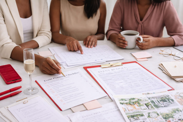 Team of women collaborating on event planning with documents, clipboard, and coffee on a styled workspace.